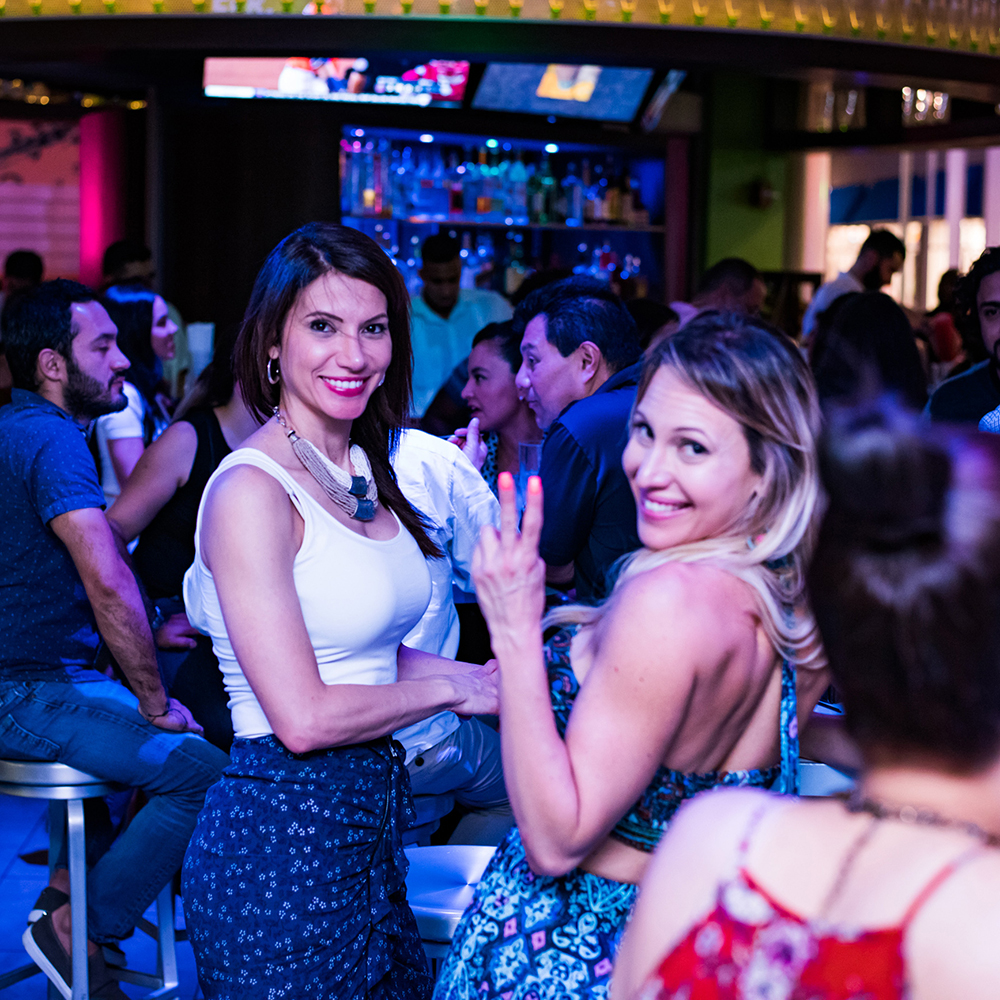 Blonde woman waves the peace sign as her friend with brown hair smiles with her at the bar