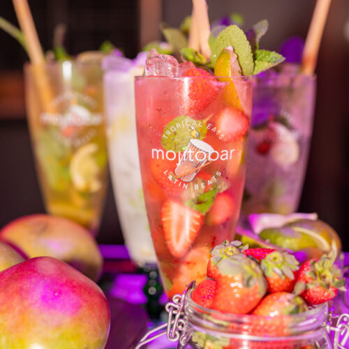 glass of strawberry mojito with several glasses in the background and fresh fruits bowl